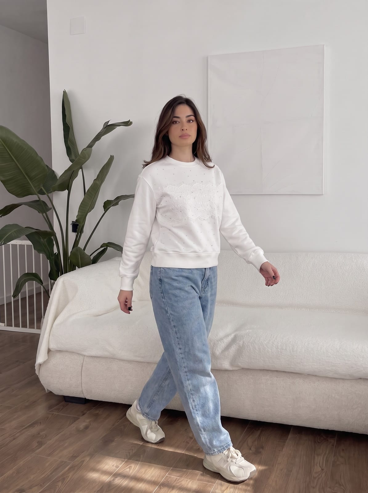 Model walking in a white studio in a white sweatshirt with lace detail and jeans.